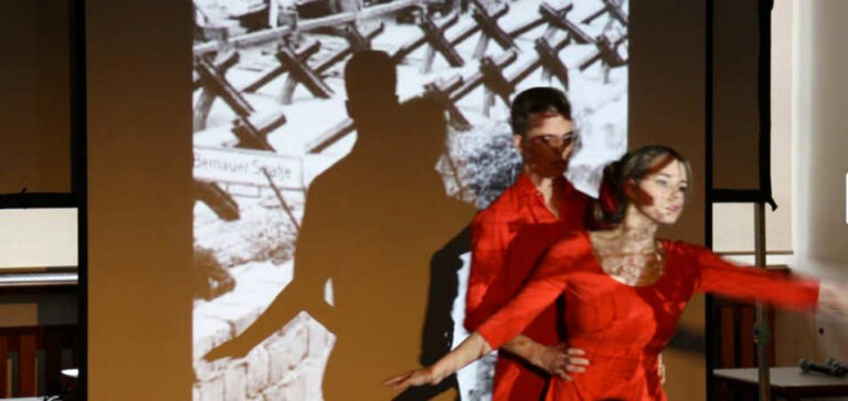 Tanz in roten Kleidern vor Schwarz-Weißaufnahmen mit Kriegsmotiven mit Musik aus der 5. Sinfonie von Beethoven